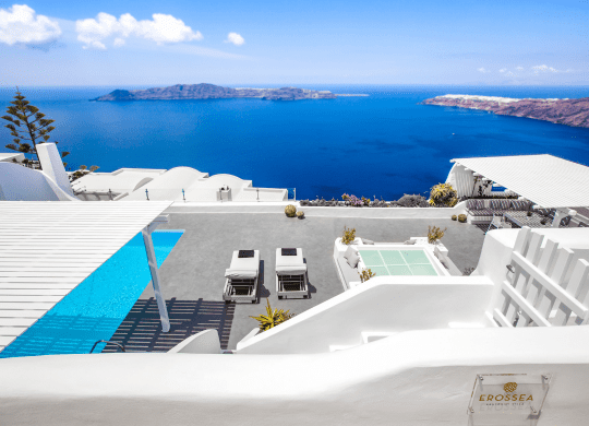 Roof top terrace at Erossea Villa featuring a bright blue pool and several sun loungers against a backdrop of white architecture. The view includes a distant landscape with mountains and the sea under a partly cloudy sky.