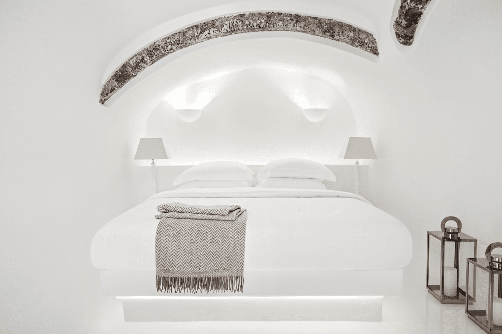 Minimalist white bedroom at Erossea Villa in Santorini with a large bed under an arched ceiling. The bed is adorned with two taupe lamps, wall sconces, and a patterned throw blanket. Two lanterns with candles are on the floor beside the bed, adding a cozy touch.