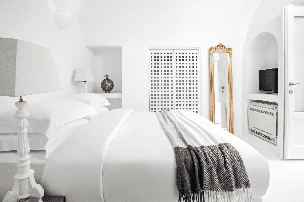 A minimalist white bedroom at Erossea Villa features a plush bed with a gray throw blanket, a white bedside lamp, and a decorative lamp in the corner. A tall gold-framed mirror stands near a TV and air conditioner by a latticed cabinet.