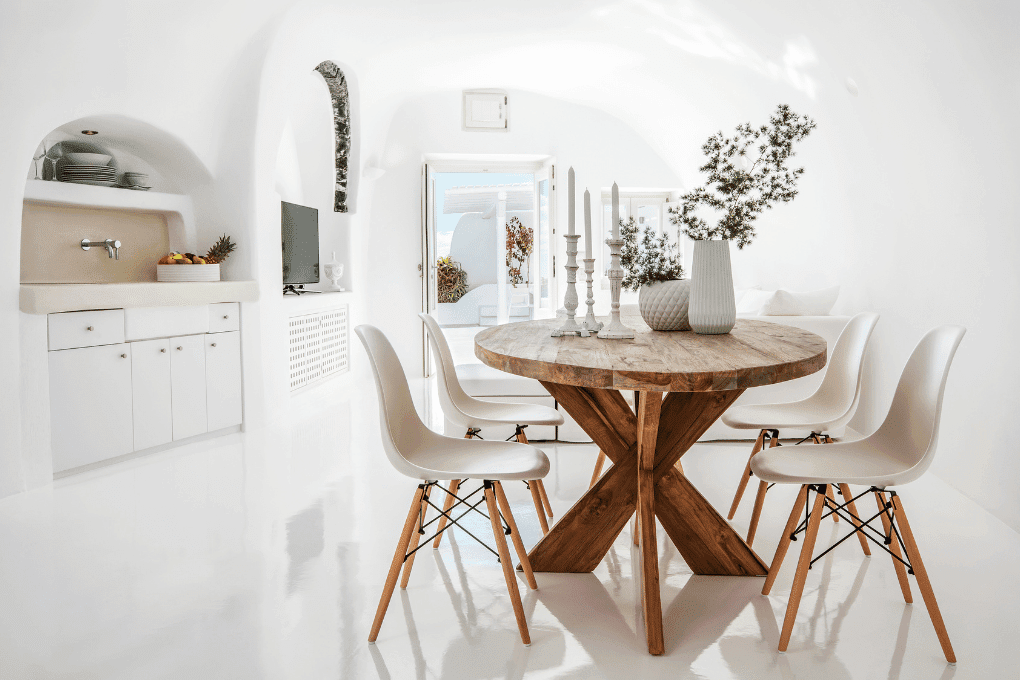 Minimalist white living room with a wooden coffee table, gray couch, and dining area. Large open window reveals a scenic view of the sea and sky. Decor includes plants and books, creating a bright, airy, and serene atmosphere.