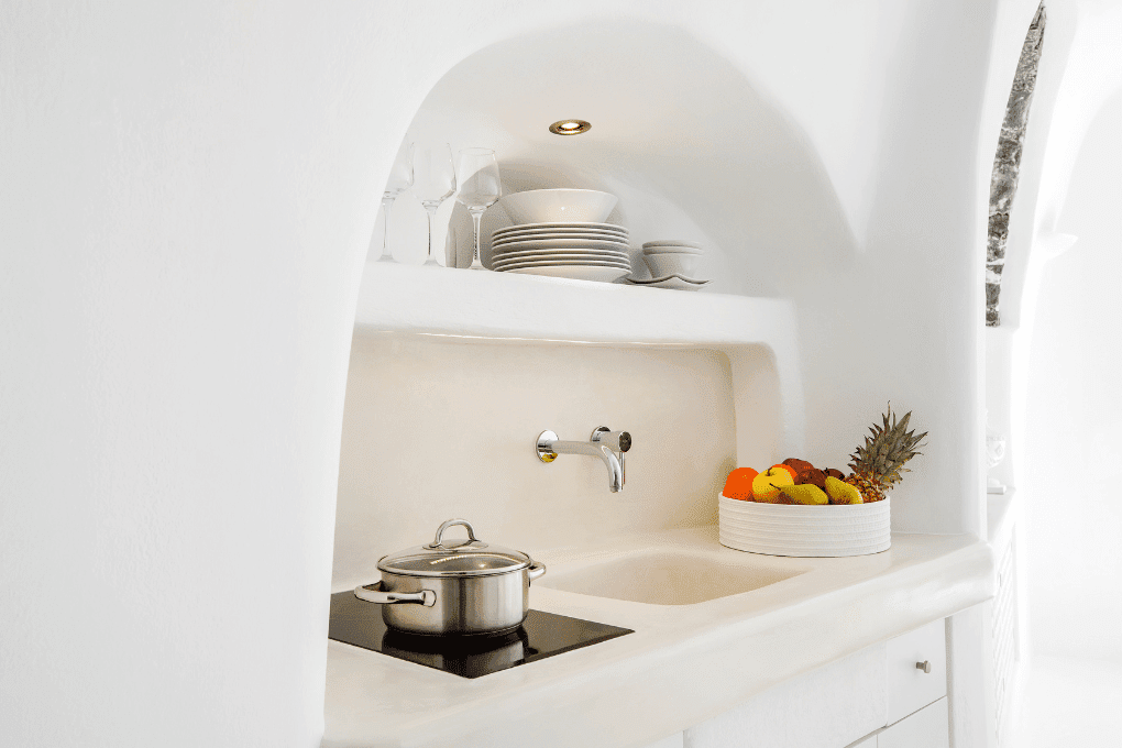 Minimalist kitchen at Erossea Villa in Santorini with white countertops and walls. Features a stovetop with a pot, a sink, and an open shelf holding plates and glasses. A bowl of mixed fruit, including apples and pineapples, is placed beside the sink.