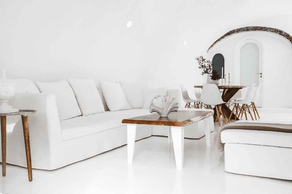 Minimalist white living room at Erossea Villa in Santorini with a white sofa and a modern wooden coffee table. In the background, theres a dining area with a wooden table and white chairs. The room features arched doorways and a bright, airy atmosphere.