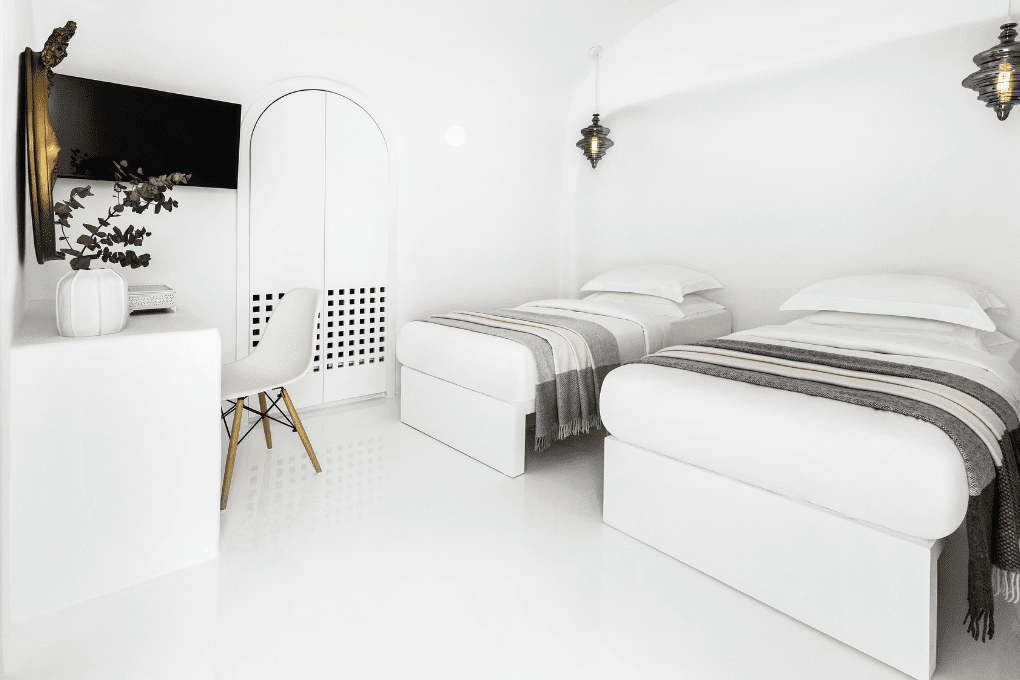 Erossea Villa minimalist, all-white room with two single beds, each with striped gray and white blankets. A wall-mounted TV, chair, and vase with branches are on the left. Modern hanging lights add a touch of elegance.