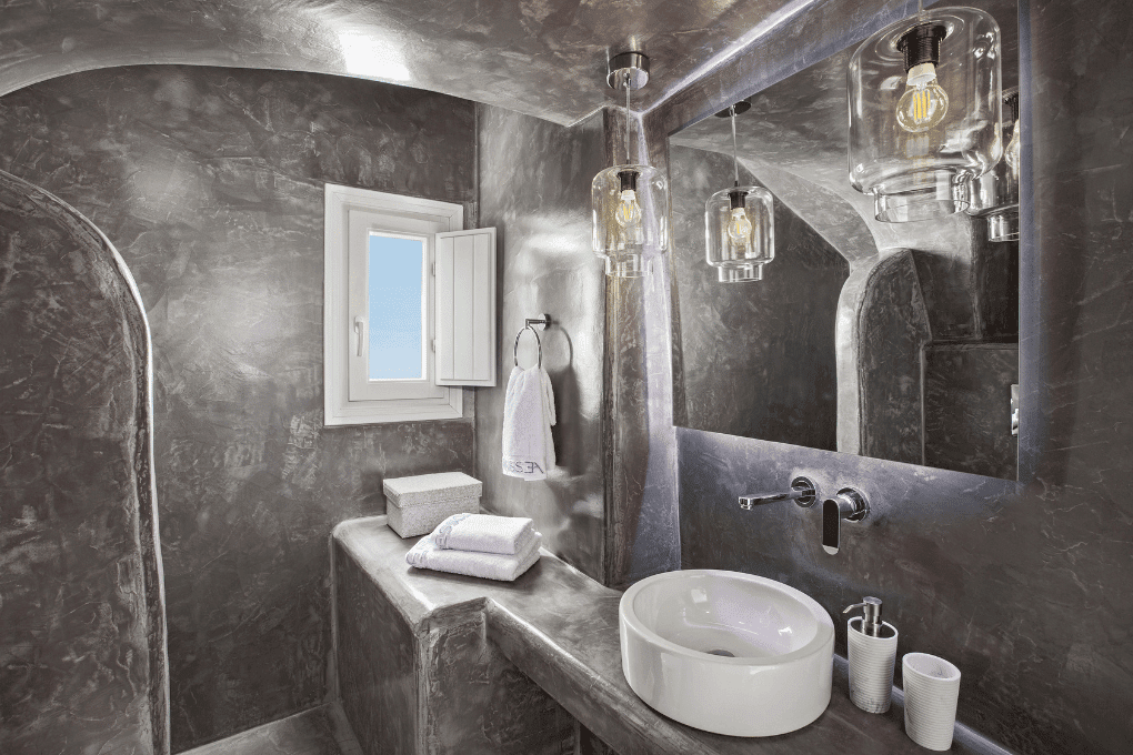 Modern bathroom in Erossea Villa with curved, concrete-like walls and an open shower. It has a built-in sink and shelves with towels. A skylight above illuminates the space, creating a minimalist and industrial feel.