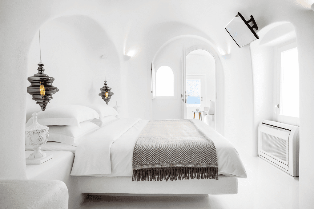 A serene, white bedroom in Erossea Villa with arched ceilings features a minimalist design. A bed with white linens and a brown throw is flanked by modern pendant lights. A wall-mounted TV faces the bed, and a door leads to a bright space beyond.