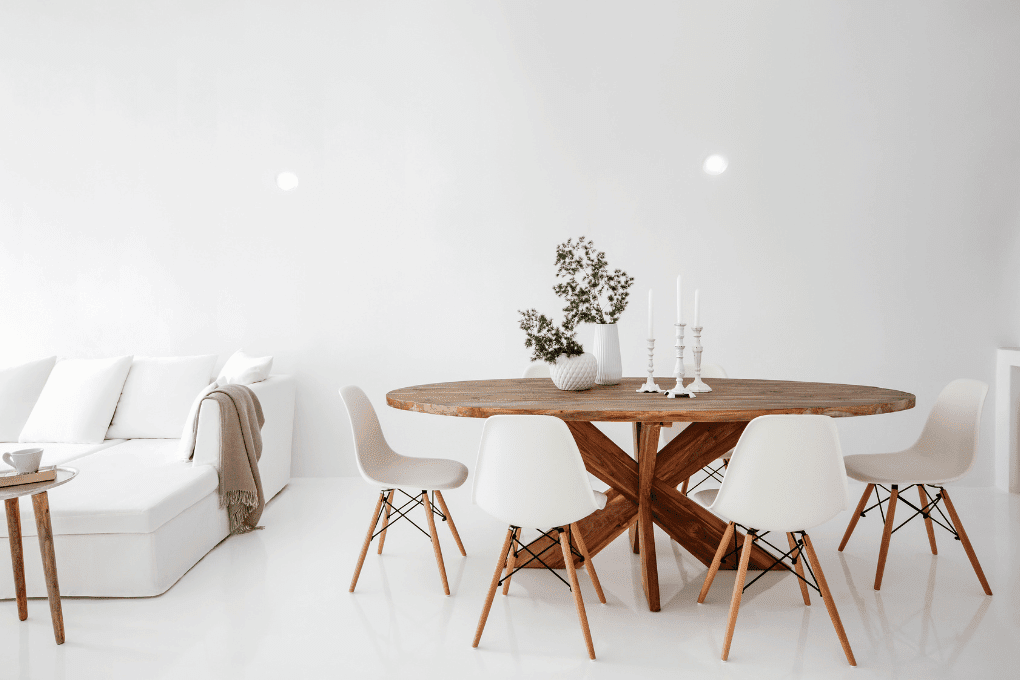 Modern dining area at Erossea Villa with a round wooden table surrounded by four white chairs. On the table, theres a ceramic vase with green foliage and a pair of white candlesticks. In the background, a white sofa is partially visible. The room is white and bright.