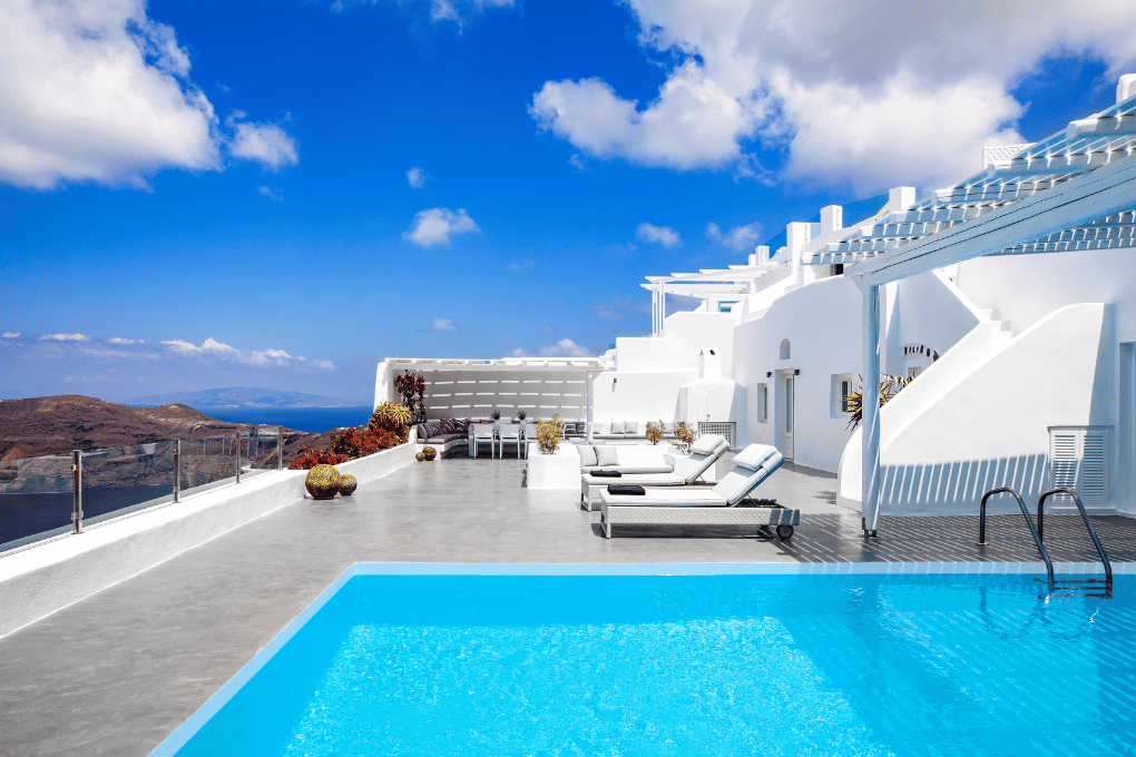 Roof top terrace at Erossea Villa featuring a bright blue pool and several sun loungers against a backdrop of white architecture. The view includes a distant landscape with mountains and the sea under a partly cloudy sky.