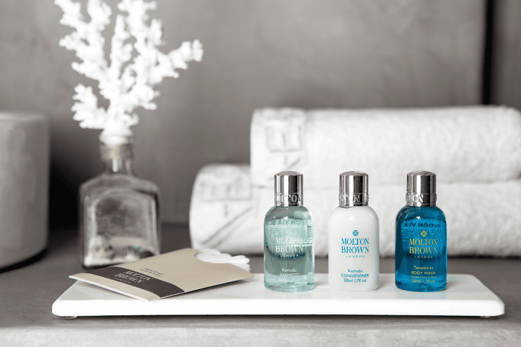 A set of toiletries, including a shampoo, conditioner, and body wash, are neatly arranged on a tray at Erossea Villa in Santorini. A decorative coral piece in a glass bottle and folded white towels are in the background, creating a luxurious bathroom setting.