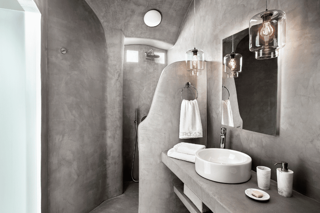 Modern bathroom in Erossea Villa with curved, concrete-like walls and an open shower. It has a built-in sink and shelves with towels. A skylight above illuminates the space, creating a minimalist and industrial feel.