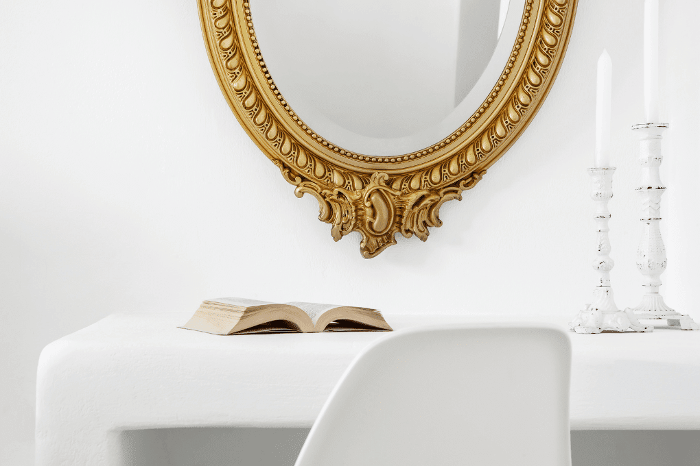 A white desk with an open book sits beneath an ornate oval mirror with a gold frame mounted on the wall. Two tall white candlesticks are on the desk. A modern white chair is partially visible in the foreground.