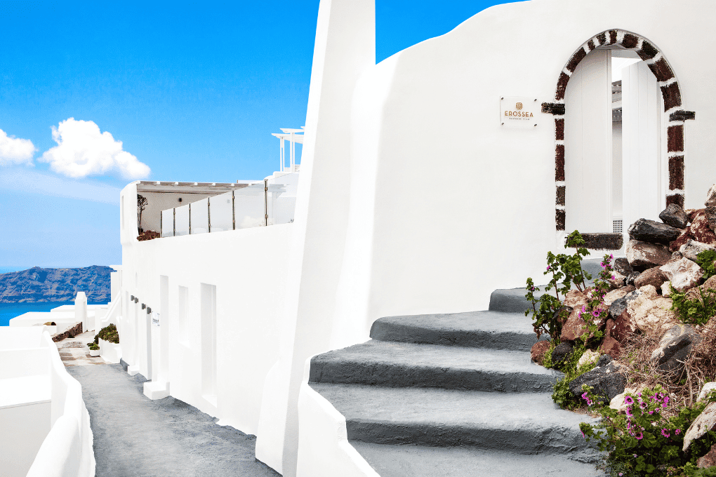 Whitewashed buildings of Erossea Villa with curved arches overlook a narrow path in Santorini. Stone steps lead to a doorway framed in dark stone. The bright blue sky and distant sea enhance the scenic view.