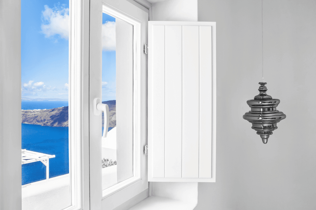 White interior of Erossea Villa with a window partially open, revealing a view of a bright blue sea and distant Santorini mountains. A modern, metallic hanging lamp is on the right side against a plain white wall.