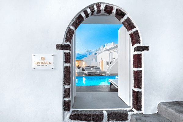 View through an arched doorway reveals a luxurious outdoor pool area with white Santorini buildings The Erossea Villa sign is mounted on the white wall.