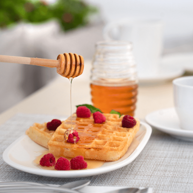 A plate of waffles topped with raspberries is being drizzled with honey from a wooden dipper. Theres a jar of honey in the background, along with a white cup and a partially visible plant. The scene is set on a light-colored tablecloth as part of the breakfast in Erossea Villa Santorini.