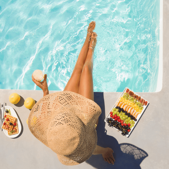A woman with a straw hat sits by a sparkling pool, legs dipped in water. Theyre holding a drink and next to them are a plate of fruit and a dish with a waffle topped with strawberries and whipped cream. Lemons are scattered nearby.