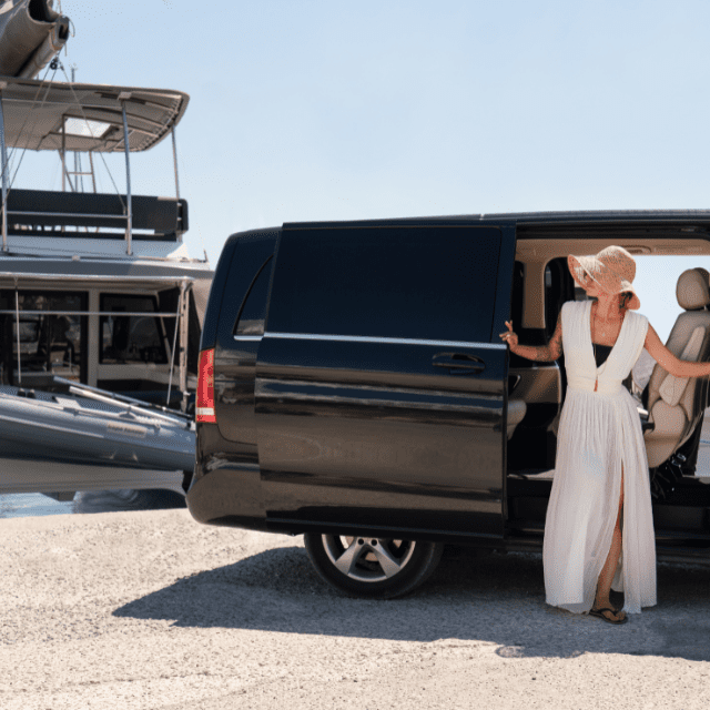 A person wearing a white dress, sunglasses, and a hat exits a black van parked near a marina. A yacht is visible in the background on a sunny day.