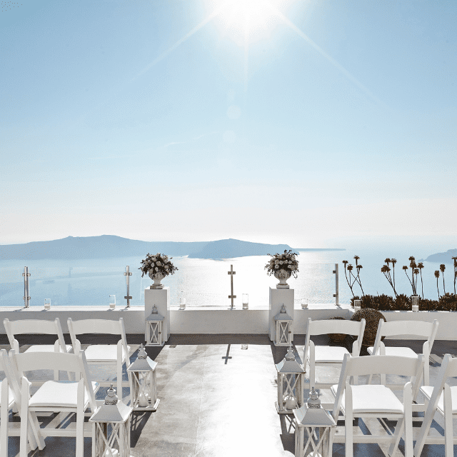 Outdoor wedding setup on the Erossea Villa terrace overlooking the sea, with white chairs and lanterns lining the aisle. Floral arrangements are placed on pillars, and a cross stands at the end. The sun shines brightly in a clear Santorini sky over the distant water.