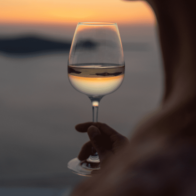 A person holding a wine glass at sunset, with a blurred landscape in the background. The golden light reflects through the glass, creating a warm and serene atmosphere.
