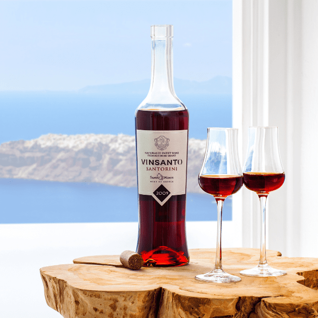 A bottle of Vinsanto wine on a wooden table with two filled wine glasses. The background features a scenic Santorini view of the blue sea and a distant island under a clear sky, visible through a window in Erossea Villa.