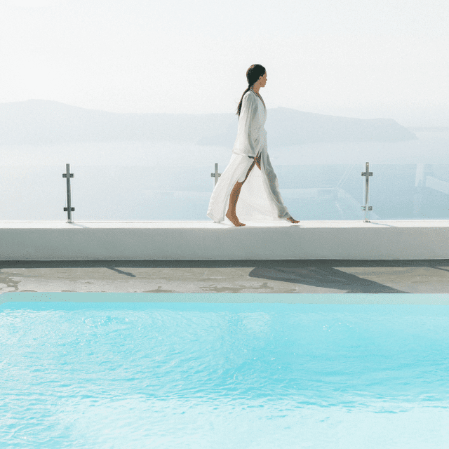 A serene woman in a flowing white robe gracefully walks along a ledge beside a tranquil pool at Erossea Villa Santorini