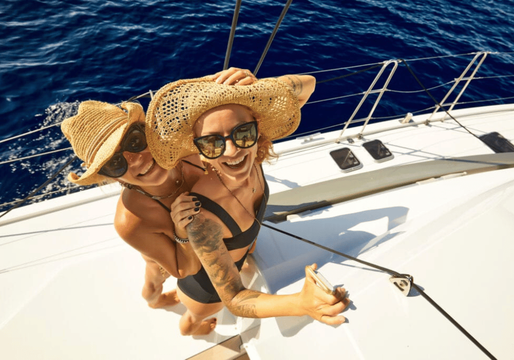 Two women wearing hats and sunglasses enjoy a sailboat ride, under a clear blue sky