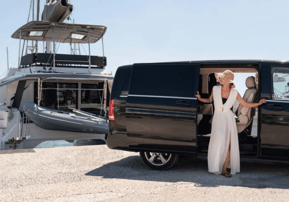 A woman wearing a white dress stands next to a black van, with the stunning scenery of Santorini in the background