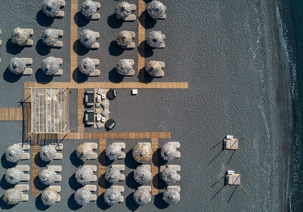 Wat Stories beach bar in Perivolos Santorini in a top down aerial view, showcasing the black sand beach and sun beds